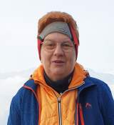 Porträt einer lächelnden älteren Person mit roten Haaren, die eine orange Jacke, eine blaue Outdoor-Jacke und ein graues Stirnband trägt, vor einem bergigen Hintergrund.