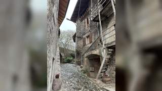Enge Kopfsteinpflastergasse mit alten Stein- und Holzgebäuden, dekoriert mit Blumen in Töpfen und hängenden Körben.