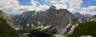 Panoramablick auf hohe, schroffe Berggipfel mit vereinzelten Schneefeldern unter einem teils bewölkten blauen Himmel, davor grüne, felsige Hänge und ein bewaldetes Tal.
