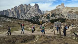 Mehrere Personen wandern auf einem Bergpfad in alpiner Landschaft, im Hintergrund steile Felsgipfel und blauer Himmel.