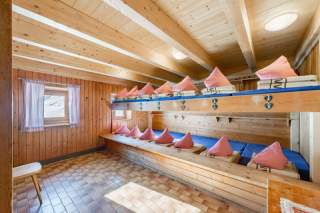 Holzsauna mit zwei Sitzreihen, ausgestattet mit blauen Sitzpolstern und pinken Kopfstützen, hellem Holzboden und einem Fenster mit Gardine.