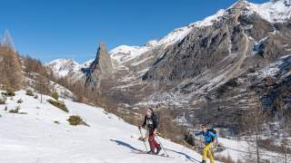 Zwei Skitourengeher mit Rucksäcken und Skistöcken steigen bei sonnigem Wetter einen schneebedeckten Bergpfad in einer alpinen Landschaft mit schroffen Felsen und vereinzelten Bäumen hinauf.
