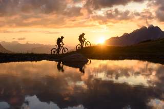Zwei Mountainbiker*innen bei untergehender Sonne, ein See im Vordergrund, das Bergpanorama im Hintergrund.